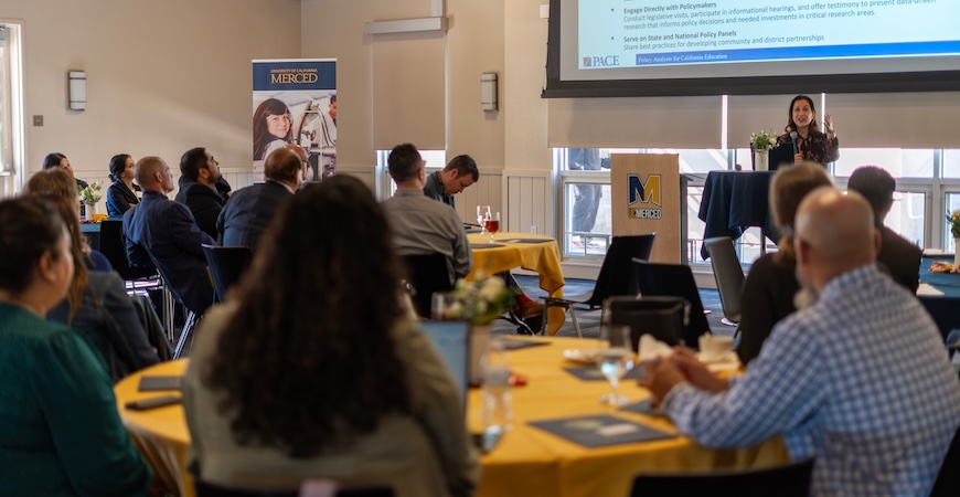 Photo depicts people sitting at tables listening to a speaker at the head of the room.